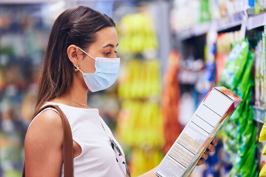 Woman reading food box information or list at the grocery store, shop or supermarket. Trendy, young female shopping in covid pandemic with a mask for organic, fresh and healthy ingredients.