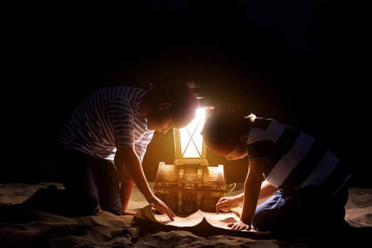 Children dressed as sailors play on the sand at night by the light of a lantern and imagine themselves as pirates looking for treasures on the map. Funny kids dream of adventure and travel.
