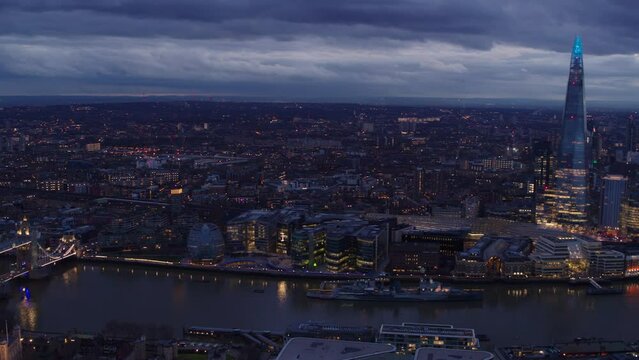 Cinematic panning aerial shot of the London financial district at dusk. Seen across the River Thames are the iconic Tower of London, HMS Belfast, London Bridge, and the Shard