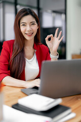 Portrait of attractive cheerful skilled girl in red suit using laptop and showing ok-sign, looking...