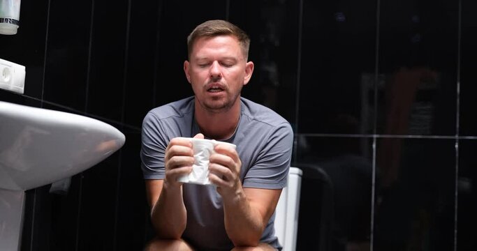 Emotional unhappy man suffering diarrhea holding roll of toilet paper sitting on toilet
