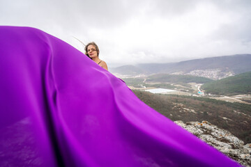 A woman with long hair is standing in a purple flowing dress with a flowing fabric. On the mountain...