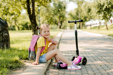 Portrait of a charming girl with a backpack and a scooter sitting on the curb in the park after school