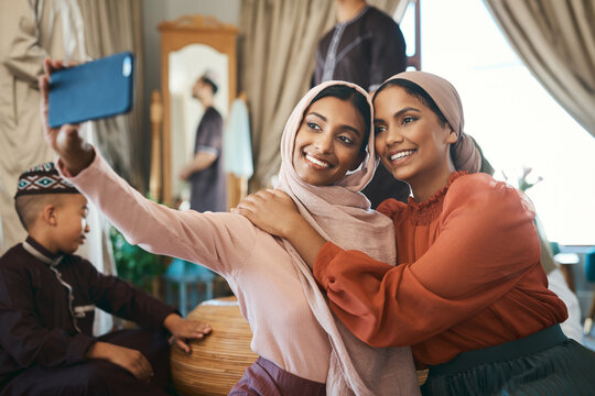 Muslim sisters taking selfies in traditional Islamic head scarf inside a happy family home together. Beautiful young women who are proud of their religion post photos of Eid Ramadan on social media