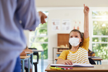 Smart student with covid face mask asking teacher question about corona virus pandemic in a classroom or elementary school. Little girl child raising hand to answer healthcare related topic in class