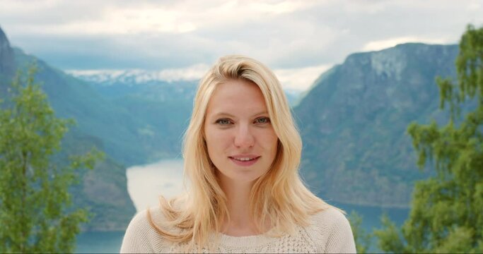 Calm, satisfied nature activist outdoors by a mountain and green trees in a beautiful landscape. Portrait of a young Norwegian blonde woman or tourists relaxing outside by a lake or river