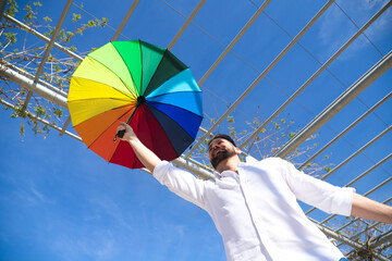 Handsome young gay man with blue eyes and white shirt stretches out his arm with a rainbow coloured umbrella in his hand. The photo is taken from below and the blue sky can be seen in the background.