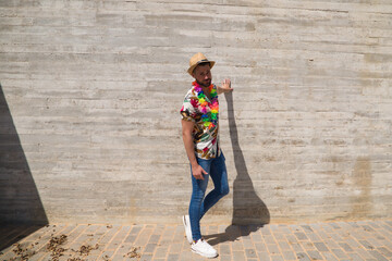 Handsome young man wearing a shirt with Hawaiian flowers, a hat and a necklace with coloured flowers. The man is happy. Background grey wall. Travel and holiday concept.