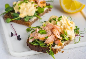 Smorrebrods, open Danish sandwiches with baked salmon, egg salad and microgreens on a white ceramic board against a light concrete background. Sandwich recipes.