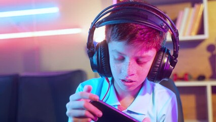 Serious teenager kid with headset playing video game on mobile phone at home - concept of relaxation, technology addiction and entertainment