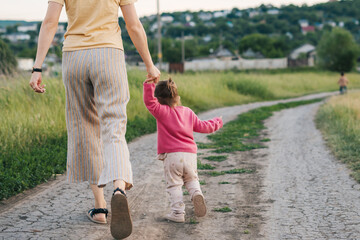 Fototapeta premium Back view of a beautiful young mother with daughter holding hands walking on a country road. Back view. Happy family, childhood. Parent, child. Mother nature.