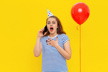 Surprised young woman is talking on a mobile phone, receives congratulations from friends on anniversary, wears party cone and striped t shirt, feels very happy, standing over yellow background