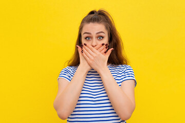 Scared girl in white-blue striped t shirt covers mouth hands and looking at camera with shocked facial expression. Young emotional woman