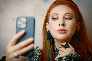 Red-haired woman takes a selfie on the front camera of a modern smartphone. Female blogger films herself for a fashion and makeup blog. Cute girl with jewelry in Gothic style. Witch aesthetic.