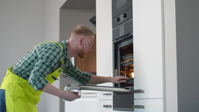 Professional worker in uniform checking broken oven to find defect in spacious kitchen. Realtime