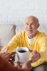 Smiling granddad looking at blurred african american grandson with cup of tea at home.