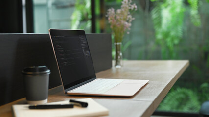Laptop with programming code on screen, coffee cup and notebook on wooden office desk