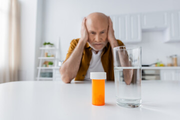 Pills and glass of water near blurred pensioner at home.