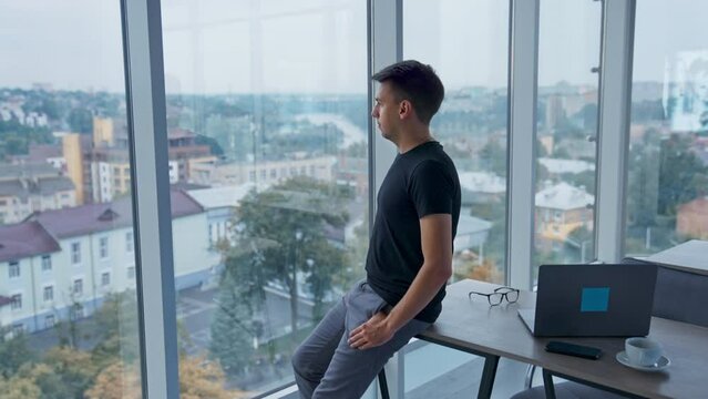 Approaching the young man sitting on the desk in office. Male looking into window and then turns to the camera smiling. Blurred backdrop.