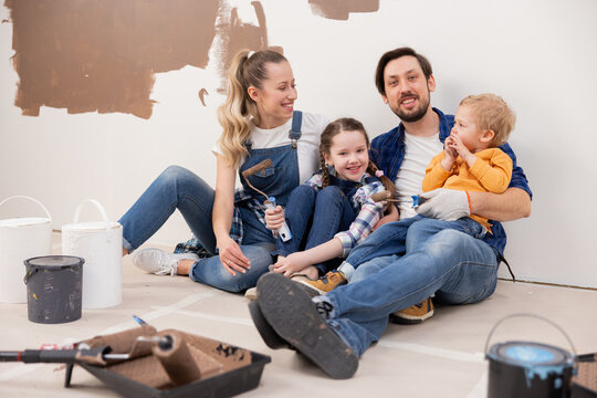 family dressed in denim style sitting on the floor. Repairs are taking place around them. They are happy about the fact that they finally have the opportunity to repaint the walls in the living room.