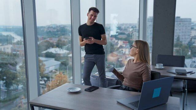 Young colleagues talking in the office. Lady sitting at desk drinking coffee. Office with panoramic windows at backdrop.