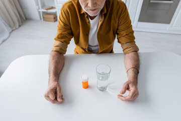 Cropped view of pensioner siting near pills and glass of water in kitchen.