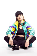 Closeup Portrait of Young Caucasian Female  Sportwoman in Fitness Outfit and baseball Cap With Wireless Headphones Listening Music While Sitting On Floor With Legs Crossed Over White.