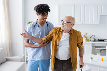 Obraz premium Positive african american nurse in uniform looking at pensioner with walking cane at home.