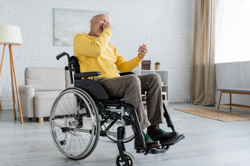Laughing senior man holding smartphone while sitting in wheelchair at home.