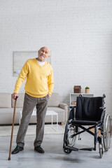 Fototapeta na wymiar Senior man with walking cane standing near wheelchair at home.