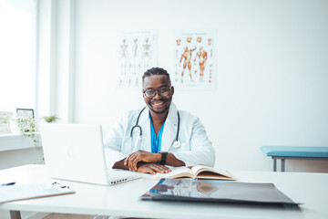 Portrait Of Smiling male Doctor Wearing White Coat With Stethoscope Sitting Behind Desk In Office....