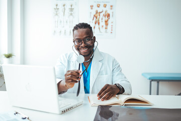 Doctor in office working on laptop. Male doctor working at office desk and smiling at camera, office interior on background. Portrait of doctor in eyeglasses and in white coat sitting at his workplace
