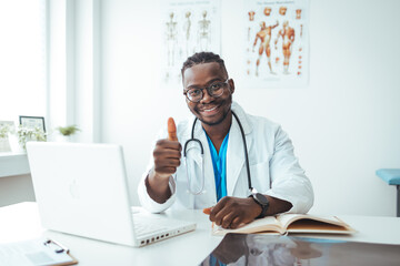 Young handsome doctor man wearing white profressional coat in office doing happy thumbs up gesture with hand. Approving expression looking at the camera showing success. 