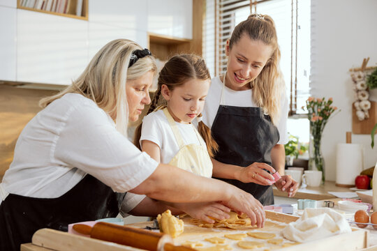 Adorable young girl wearing apron pressing cookies with cookies cutter helping granny and mother cooking preparing homemade domestic cookies biscuits.
