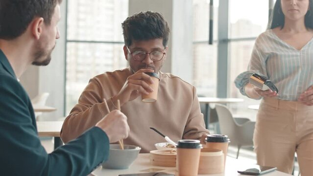 Waist up slowmo of young Biracial man in smart casualwear and eyeglasses paying for lunch with colleague with smartphone, sitting at table on modern food court with panoramic windows