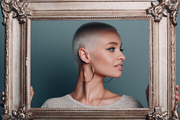 Millenial young woman with short blonde hair holds gilded picture frame in hands behind her face...