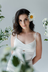 Portrait of young model with chamomile in hair looking at camera near flowers isolated on grey.