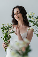 Pretty brunette woman in white top touching flowers isolated on grey.