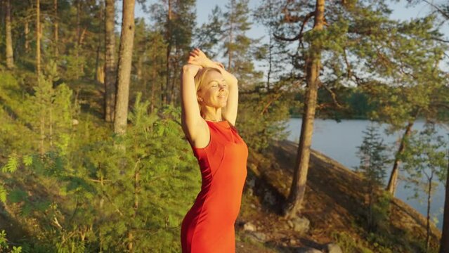 A beautiful woman in a long red dress stands near a forest lake at sunset.