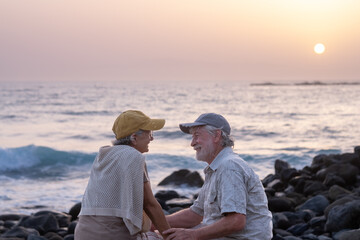 Portrait of two happy and youthful seniors or retirees sitting on the beach in sunset light hands...