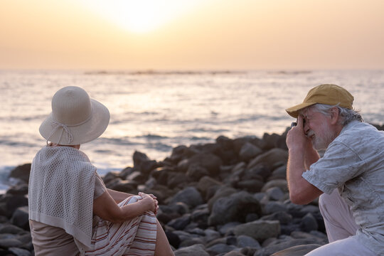 Senior man takes a picture to his wife sitting on the beach at sunset light