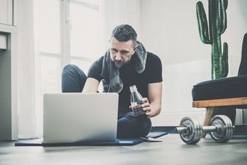 Sporty man resting after working out at home. Fit boy looking gym exercise video on laptop computer. Strong male do online gymnastics lesson in the living room. Wellness health sport, training concept