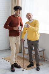 Cheerful interracial granddad and grandson holding cups and looking at camera at home.