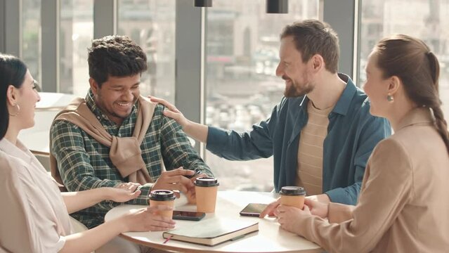 Waist up slowmo of team of multiethnic colleagues sitting at cafe table by panoramic window, having conversation during coffee break