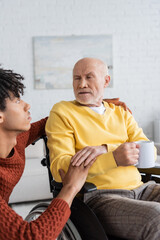 Fototapeta premium African american grandson touching hand of granddad with cup in wheelchair at home.