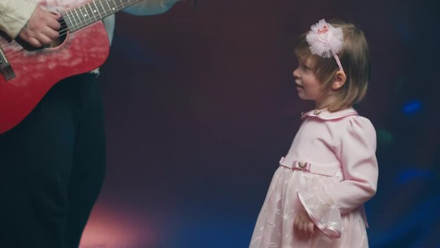 Little girl on stage in pink vintage dress, she dances, her father plays acoustic guitar. Color music is shining. Performance on stage. Early development of children