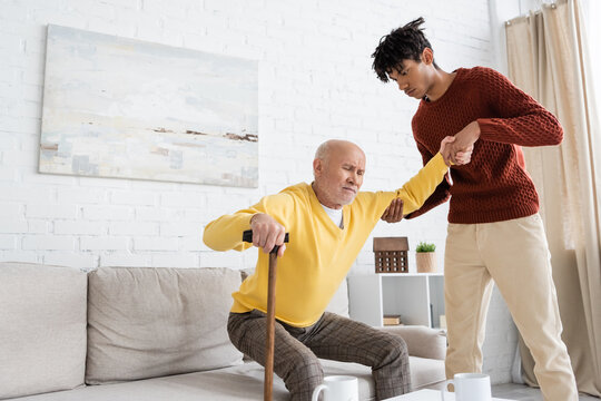 African american grandson helping displeased granddad with walking cane at home.