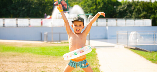 A cheerful child boy in bright turquoise and orange swimming trunks stands with an inflatable ring against the background of a summer street water park