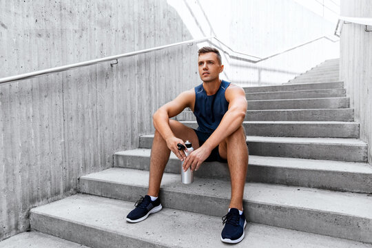 fitness, sport and people concept - tired young man with bottle of water sitting on stairs