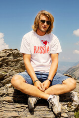 a long-haired young man with a beard and glasses in a white T-shirt with the inscription Russia on the background of mountains